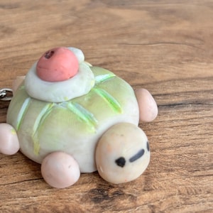 May include: A small, handmade clay turtle figurine featuring a pale green shell with light green accents, a smooth off-white head with two black dot eyes, and four rounded light pink limbs. A white dollop with a pink berry sits on the shell.