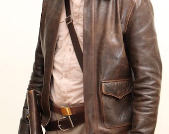 May include: A brown leather jacket with a collared neckline and two front pockets. The jacket is worn over a light pink button-down shirt and tan pants. A brown leather belt with a gold buckle and a brown leather shoulder bag complete the look. A whip is held in the left hand.