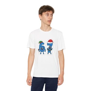 May include: White t-shirt featuring a cartoon design of the numbers 6 and 7, each with a face and festive holiday hats. The number 6 wears an elf hat, and the number 7 wears a Santa hat. The numbers are blue with black outlines.