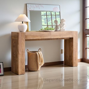 May include: A light brown wooden console table with a rectangular mirror, a white lamp, and decorative items. A woven basket with a beige throw is placed beneath the table. The table is made of solid wood and has a natural finish.