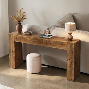 May include: A rustic wooden console table with a natural finish, featuring a rectangular top and two thick legs. The table is adorned with a vase of dried flowers, a small lamp, a book, and a decorative bowl. A small, round, upholstered stool sits below.