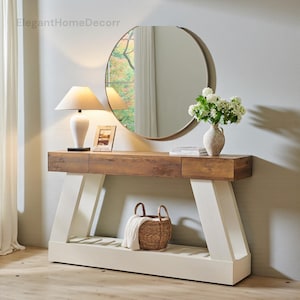 May include: A wooden console table with a light brown top and white legs. A round mirror hangs above the table, which is decorated with a lamp, a framed photo, a vase of flowers, and a woven basket.