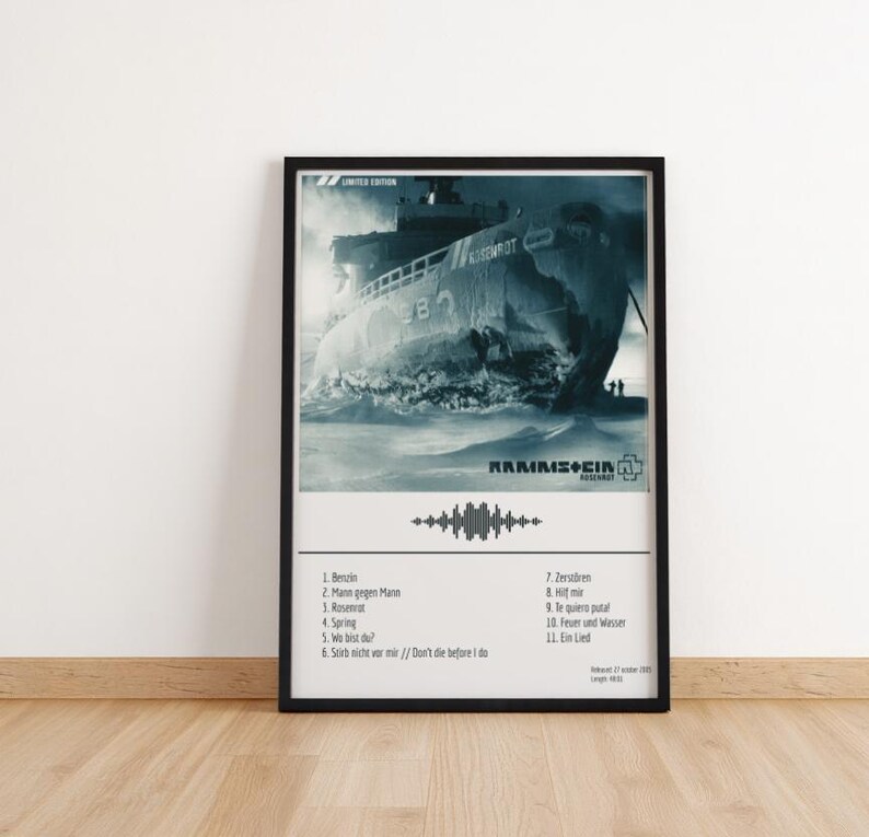 May include: A framed poster featuring a grayscale image of a ship breaking through icy waters. The poster includes the band name "Rammstein" and a tracklist. The poster is in a black frame and is displayed on a wooden floor.