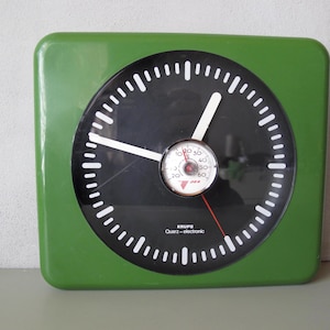 Könnte beinhalten: Eine quadratische, grüne Wanduhr mit schwarzem Zifferblatt und weißen Markierungen. Die Uhr hat weiße Zeiger und ein kleines Messgerät in der Mitte. Der Text "KRUPS Quartz-electronic" ist auf dem Zifferblatt aufgedruckt.