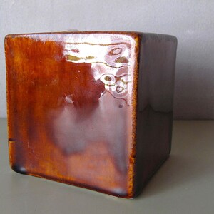 May include: A square, ceramic container with a glossy, brown glaze. The cube-shaped object has a smooth surface with a slight sheen, reflecting light. The edges are slightly darker, creating a subtle contrast. The container is likely for decorative or functional use.