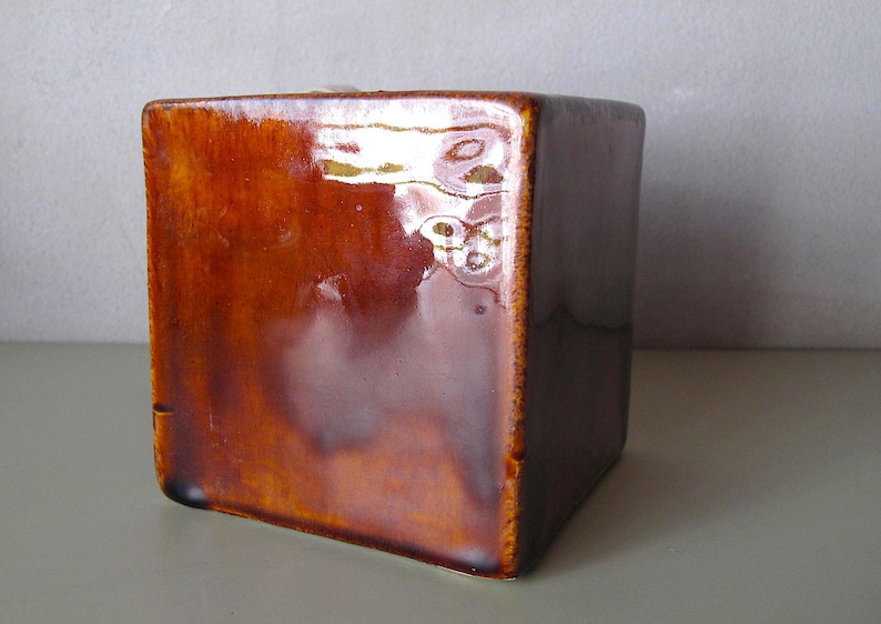May include: A square, ceramic container with a glossy, brown glaze. The cube-shaped object has a smooth surface with a slight sheen, reflecting light. The edges are slightly darker, creating a subtle contrast. The container is likely for decorative or functional use.