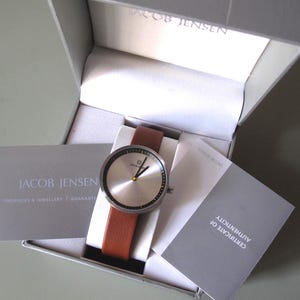 May include: A silver wristwatch with a brown leather strap, displayed in a grey box. The watch face is circular with black markings and yellow hands. The box contains cards with the text "JACOB JENSEN".