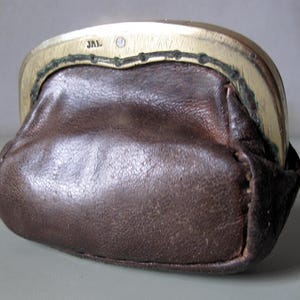 Könnte beinhalten: Eine kleine, braune Leder-Geldbörse mit einem goldfarbenen Metallrahmen. Die Geldbörse hat eine abgerundete Form und eine strukturierte Oberfläche. Der Rahmen ist mit den Buchstaben "JAL" markiert.