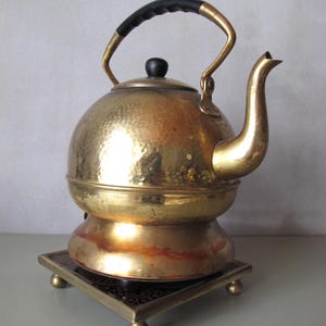 Op de afbeelding: Een vintage messing ketel met een zwarte handgreep en een decoratieve basis. De ketel heeft een afgeronde body en een gebogen tuit. De basis is vierkant met decoratieve voetjes. De ketel wordt waarschijnlijk gebruikt om water te koken voor thee of koffie.