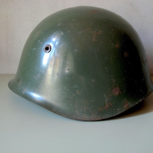 Könnte beinhalten: Ein olivgrüner Militärhelm mit abgerundeter Form. Der Helm hat ein kleines Loch an der Seite und weist Gebrauchsspuren auf, darunter Rostflecken. Die Oberfläche ist glänzend und reflektiert das Licht.