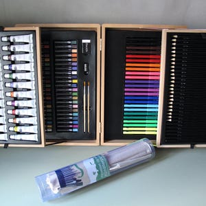 Può includere: Un set artistico in legno aperto, contenente tubetti di vernice, matite colorate e pastelli. Una custodia di plastica trasparente con pennelli e altri materiali artistici è in primo piano. Il set mostra una varietà di materiali artistici.