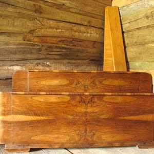 Puede incluir: Un marco de cama de madera vintage con un acabado marrón cálido y rico. El cabecero y el piecero presentan un diseño curvo y patrones intrincados de vetas de madera. El marco de la cama está colocado contra una pared de madera rústica.