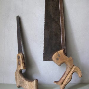 Pode incluir: Duas serras manuais antigas com cabos de madeira. Uma serra é menor com um cabo curvo, e a outra é maior com um cabo ornamentado. Ambas as serras têm lâminas escuras e envelhecidas com dentes visíveis.