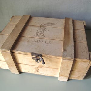 May include: A light-coloured wooden crate with the word "SAMPLES" printed on the lid. The crate is secured with wooden straps and a metal clasp. The crate has a vintage, rustic appearance.