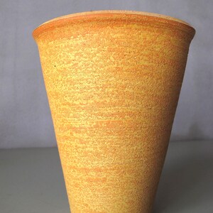 Może przedstawiać: Teksturowany, żółto-pomarańczowy ceramiczny wazon. Wazon ma cylindryczny kształt z lekko rozszerzaną górą. Powierzchnia ma nakrapiany wygląd, a obręcz jest ciemniejsza pomarańczowa.