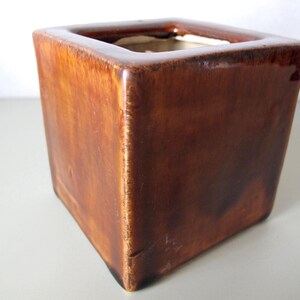 May include: A square, brown ceramic planter with a glossy finish. The planter has a cube shape with a hollow interior. The exterior has a gradient of brown tones, creating a rustic appearance. The planter is suitable for indoor or outdoor use.