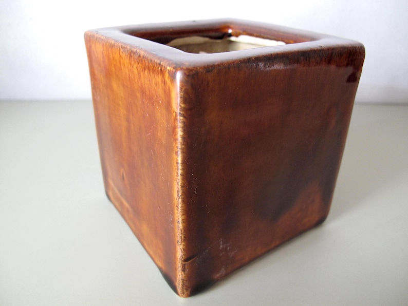 May include: A square, brown ceramic planter with a glossy finish. The planter has a cube shape with a hollow interior. The exterior has a gradient of brown tones, creating a rustic appearance. The planter is suitable for indoor or outdoor use.