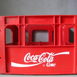 Könnte beinhalten: Eine leuchtend rote Getränkekiste aus Kunststoff mit dem weißen Coca-Cola-Logo und dem Wort Coke auf der Vorderseite. Der rechteckige Behälter hat eine gitterartige Struktur mit offenen Seiten und einen verstärkten Rahmen für Flaschen.