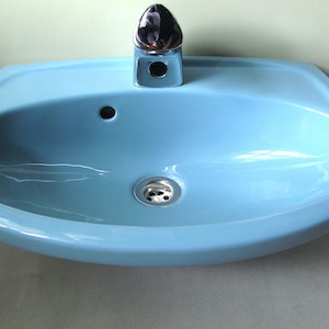 Könnte beinhalten: Ein hellblaues Keramik-Waschbecken mit einer verchromten Armatur. Das Waschbecken hat eine abgerundete Form mit einem Abfluss und einem einzelnen Wasserhahnloch. Die Armatur hat eine glänzende Chromoberfläche. Das Waschbecken ist hellblau.