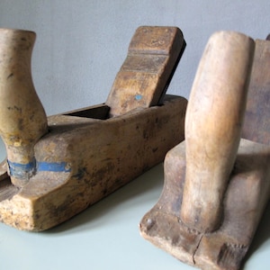 Könnte beinhalten: Zwei antike Handhobel aus Holz. Die Hobel bestehen aus gealtertem Holz mit einem gebogenen Griff und einer rechteckigen Basis. Ein Hobel hat einen blauen Akzent. Die Hobel werden wahrscheinlich für die Holzbearbeitung verwendet.