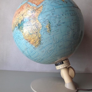 Op de afbeelding: Een vintage wereldbol met een lichtblauwe oceaan en bruine continenten. De wereldbol is gemonteerd op een crèmekleurige standaard met een cirkelvormige basis en een gradenschaal. Een netsnoer komt uit de basis.