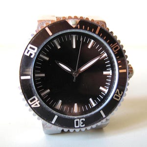 Könnte beinhalten: Eine Armbanduhr mit schwarzem Zifferblatt, silberner Lünette und einem schwarzen, drehbaren Außenring. Die Uhr hat weiße Stundenmarkierungen und Zeiger. Die Lünette ist mit weißen Ziffern in 10-Minuten-Abständen markiert. Klassisches Design.