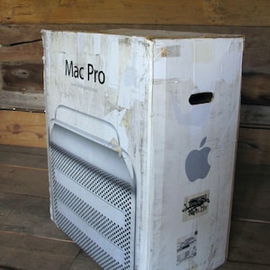 Op de afbeelding: Een verweerde kartonnen doos voor een Mac Pro workstation. De doos is wit met het Apple-logo en de woorden "Mac Pro" erop gedrukt. De doos heeft een handgreepuitsparing en toont een afbeelding van de computer.