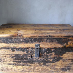 Op de afbeelding: Een verweerde houten kist met een rechthoekige vorm. De kist heeft een verweerde uitstraling met zichtbare houtnerf en donkere accenten. Een metalen sluiting zekert het deksel, geschikt voor opslag of decoratie.