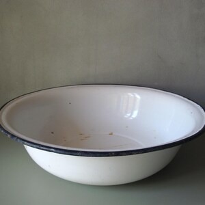 Könnte beinhalten: Eine weiße Emaille-Schüssel mit dunkelblauem Rand. Die Schüssel hat eine abgerundete Form und ein leicht abgenutztes Aussehen, was auf eine Verwendung im Vintage-Stil hindeutet. Das Innere weist leichte Gebrauchsspuren auf. Geeignet für Küche oder Dekoration.