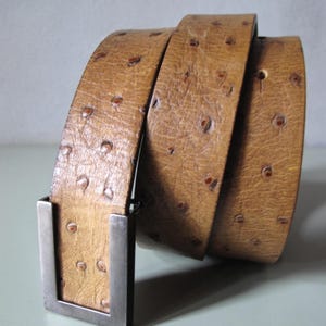 May include: A tan leather belt with a textured surface and a silver-colored rectangular buckle. The belt is rolled up, showcasing its detailed pattern and design. The belt has a series of small, dark spots.
