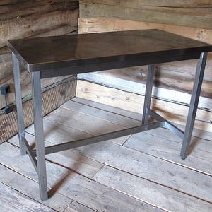 Pode incluir: Uma mesa retangular com um tampo metálico escuro e liso, e uma estrutura robusta feita de tubos de aço cinza de secção quadrada. A mesa possui quatro pernas retas conectadas por uma barra de suporte horizontal entre as pernas para estabilidade.