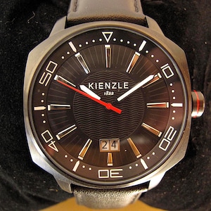 Puede incluir: Reloj Kienzle negro con correa de cuero negro. La esfera negra presenta marcadores blancos y dorados, un segundero rojo y una pantalla de fecha. La caja de acero inoxidable tiene un bisel negro con números blancos.