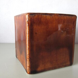 May include: A square, ceramic container with a glossy, brown finish. The sides are slightly tapered, and the corners are rounded. The container has a vintage or antique appearance, suitable for holding small items or as a decorative piece.