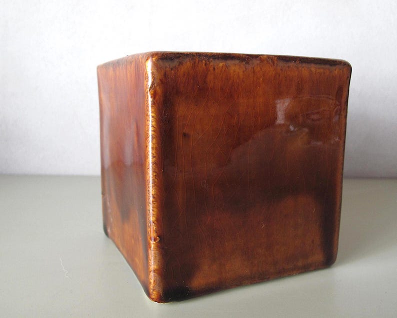 May include: A square, ceramic container with a glossy, brown finish. The sides are slightly tapered, and the corners are rounded. The container has a vintage or antique appearance, suitable for holding small items or as a decorative piece.