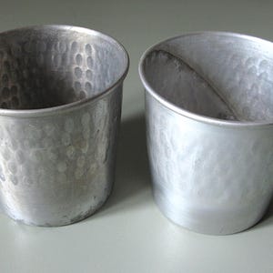 Könnte beinhalten: Zwei silberfarbene Metallbecher mit gehammerter Textur. Die Becher haben eine leicht konische Form und eine breite Öffnung. Die Becher sind leer und scheinen aus Aluminium zu sein.