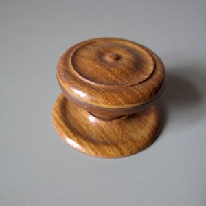 Puede incluir: Un tirador de armario de madera con una parte superior redonda y una base circular. El tirador es de color marrón cálido con vetas de madera visibles. El diseño es simple y clásico, adecuado para varios estilos de muebles.