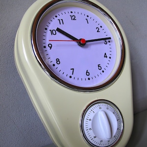 以下が含まれることがあります： クリーム色のキッチンタイマー。時計の文字盤と、別のタイマーダイヤルが付いています。時計には黒い針、赤い秒針、黒い数字があります。タイマーダイヤルは白で、黒いマークと白いノブが付いています。