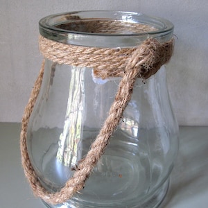 May include: A clear glass jar with a wide opening and a rounded base. The jar is wrapped with a thick, natural-colored rope around the top and has a rope handle. The jar is empty and sits on a light gray surface.