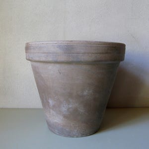 Op de afbeelding: Een grote, verweerde, bruine terracotta bloempot. De pot heeft een brede, platte rand met twee verzonken banden. De body loopt taps toe naar een kleinere basis. Het oppervlak heeft een gevlekt uiterlijk met lichtere vlekken.