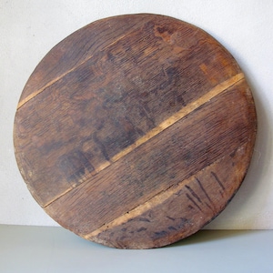 Puede incluir: Tabla de servir redonda de madera, con un aspecto rústico y envejecido. La tabla está construida con varias tablas de madera, con vetas visibles y variaciones naturales de color. La madera tiene un tono marrón cálido con vetas más claras.