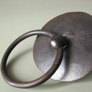 以下が含まれることがあります： テクスチャ加工された円形のバックプレートに取り付けられた、円形のハンドルが付いた、素朴な濃い茶色の金属製のドアノッカー。ノッカーは、古さと使用を暗示する風化した外観をしています。デザインはシンプルで機能的です。