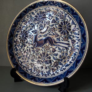 Puede incluir: Plato decorativo de cerámica con un diseño floral azul y blanco y un borde dorado. El plato presenta una imagen central de un ciervo y está sostenido por un soporte negro. El borde del plato tiene un patrón geométrico.