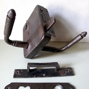 Puede incluir: Un juego de cerradura de puerta antiguo de metal marrón oscuro, con un cuerpo de cerradura rectangular con dos manijas curvas y una cerradura. Incluye una placa de manija separada y una placa de cubierta de cerradura. El juego muestra signos de edad y desgaste.