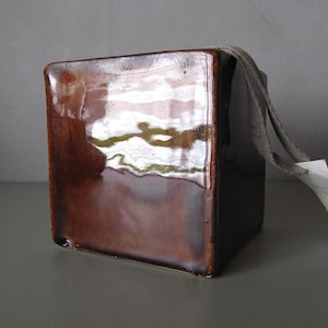 Op de afbeelding: Een vierkante, bruine keramische container met een glanzende afwerking. Een grijze stoffen lus en een wit label zijn aan de bovenkant bevestigd. De container is waarschijnlijk voor decoratieve of opslagdoeleinden.