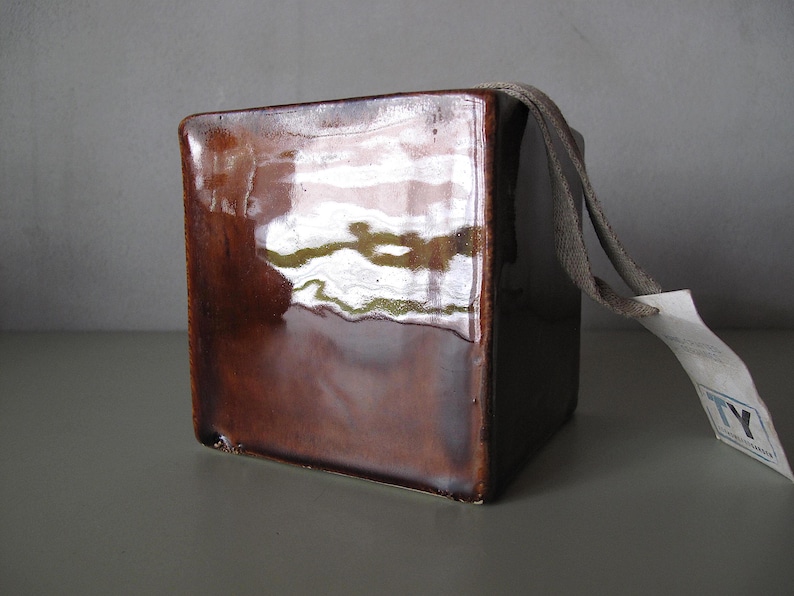 May include: A square, brown ceramic container with a glossy finish. A gray fabric loop and a white tag are attached to the top. The container is likely for decorative or storage purposes.