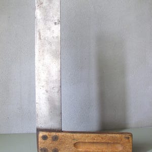 Könnte beinhalten: Ein alter Zimmermannswinkel mit Holzgriff und Metallklinge. Der Griff besteht aus hellbraunem Holz und hat eine Aussparung zum Greifen. Die Metallklinge ist silberfarben und weist Gebrauchsspuren auf. Zum Messen und Markieren von rechten Winkeln.