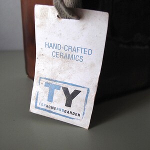 May include: A close-up of a tag attached to a ceramic container. The tag reads "HAND-CRAFTED CERAMICS" and features the letters "TY" inside a blue and black rectangle. The words "FORHOMEANDGARDEN" are below the rectangle.
