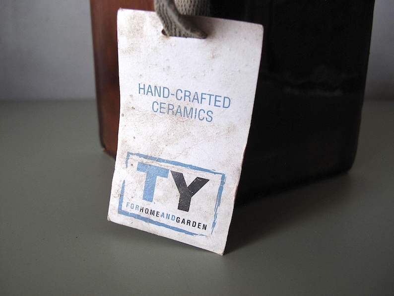 May include: A close-up of a tag attached to a ceramic container. The tag reads "HAND-CRAFTED CERAMICS" and features the letters "TY" inside a blue and black rectangle. The words "FORHOMEANDGARDEN" are below the rectangle.