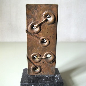 Può includere: Scultura rettangolare color bronzo con tre aperture circolari collegate da linee curve. Montata su una base di marmo nero. L'opera d'arte presenta una superficie strutturata e un design astratto unico. Altezza di circa 15 cm.
