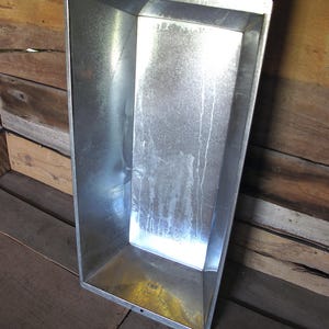 Könnte beinhalten: Ein rechteckiger Trog aus verzinktem Metall mit einer breiten, offenen Oberseite und sich verjüngenden Seiten. Die Innenfläche weist eine gesprenkelte, metallisch silberne Oberfläche mit sichtbaren vertikalen Streifen auf.